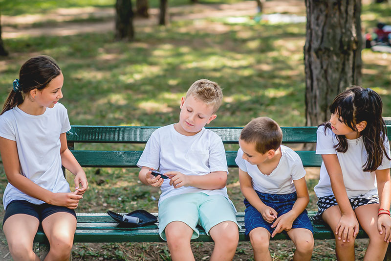 Symbolbild: Ein Junge auf einer Parkbank testet seinen Blutzuckerspiegel, drei andere Kinder schauen freundlich und interessiert zu Symbolbild: Ein Junge auf einer Parkbank testet seinen Blutzuckerspiegel, drei andere Kinder schauen freundlich und interessiert zu
