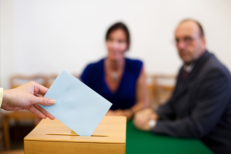 Schmuckbild: EIne Frauenhand steckt einen Wahlumschlag in eine Wahlurne, im Hintergrund unscharf ein Wahlhelfer und eine Wahlhelferin (QUelle: Adobe Stock)