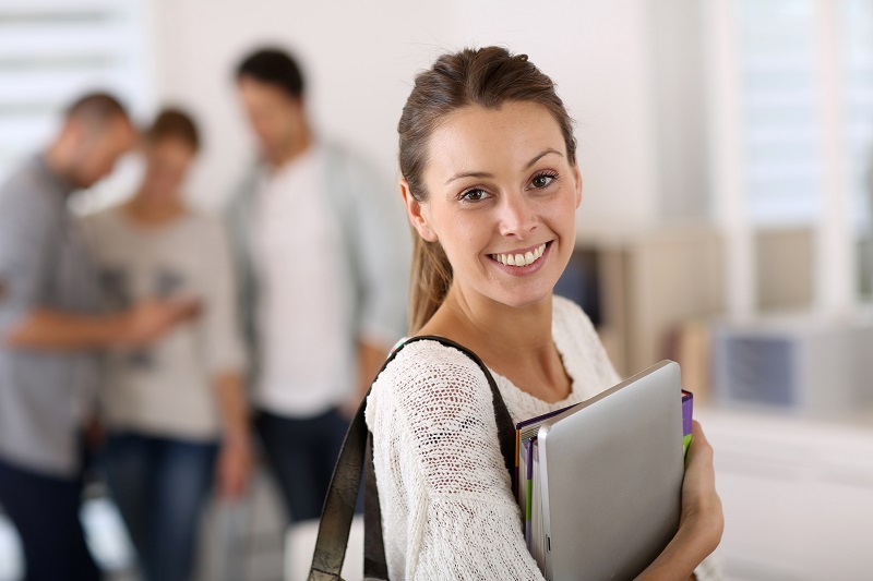 Schmuckbild: Junge Frau mit Büchern und Tablet-Computer, im Hintergrund verschwommen drei junge Menschen, die auf ein Smartphone blicken (Quelle: www.fotolia.com, von goodluz, #57959230)