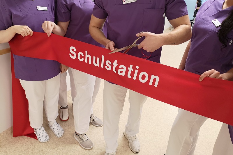 Eröffnung der Schulstation: Pflege-Auszubildende halten und zerschneiden ein breites rotes Band Eröffnung der Schulstation: Pflege-Auszubildende halten und zerschneiden ein breites rotes Band