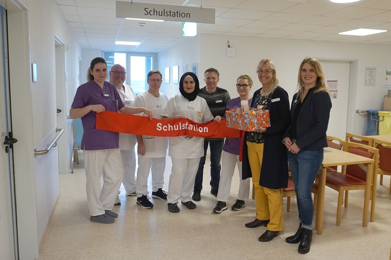 Mitarbeiter aus der Schulstation und der Krankenpflegeschule halten ein rotes Stoffband mit der Aufschrift Schulstation, eine Person hält ein bunt eingepacktes Geschenk