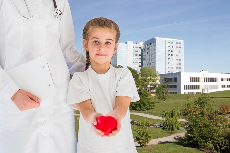Schmuckbild; ein Kind hält ein rotes Herz mit beiden Händen in die Kamera, neben ihr der Rumpf einer Person im weißen Kittel; im Hintergrund das Klinikum Schmuckbild; ein Kind hält ein rotes Herz mit beiden Händen in die Kamera, neben ihr der Rumpf einer Person im weißen Kittel; im Hintergrund das Klinikum